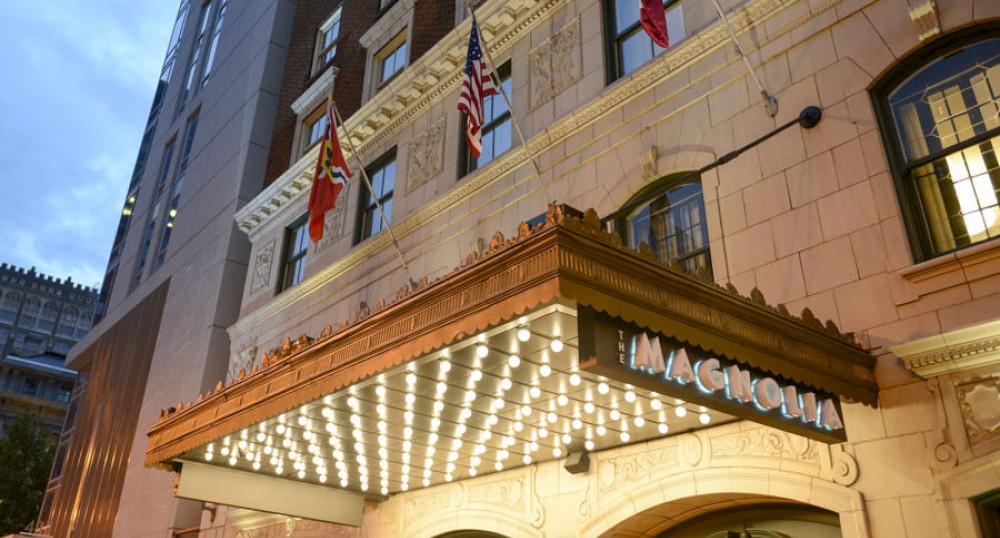 The image shows the entrance of the Magnolia Hotel with a lit marquee sign and several flags, including the American flag, displayed above.