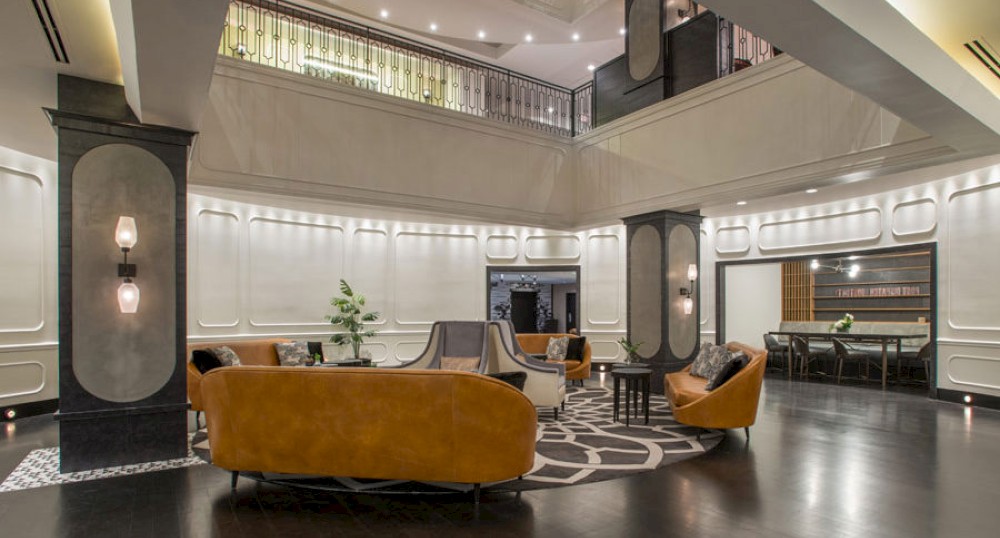A modern lobby area with mustard-colored chairs, a central seating arrangement, dark flooring, elegant lighting, and a mezzanine with railing above.