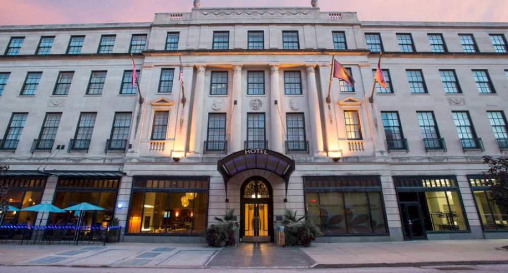 The image shows a large, multi-story hotel building with an arched entrance, several flags, and outdoor seating areas illuminated by warm lighting.
