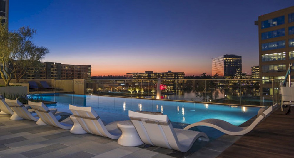 The image shows a luxurious rooftop pool area at sunset, featuring modern lounge chairs and a cityscape with illuminated buildings in the background.