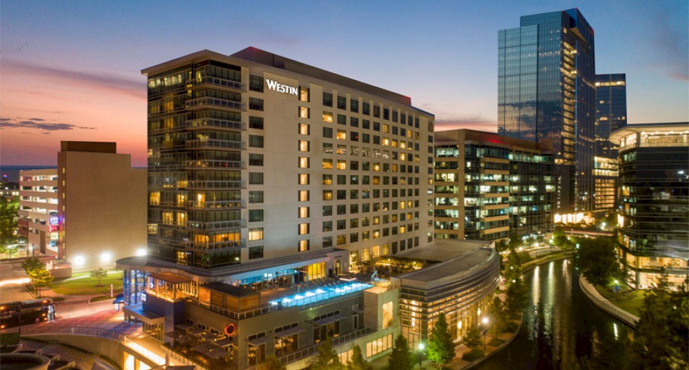 A cityscape during dusk with modern buildings, a canal, and a hotel with a rooftop pool. The sky displays a mix of colors from the sunset.