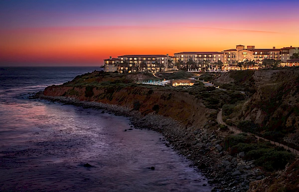 A coastal hotel is illuminated at dusk on a cliffside, overlooking a rocky shoreline with the ocean and twilight sky in the background.