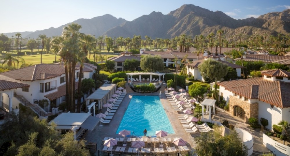 The image shows a resort with a large pool, surrounded by sun loungers and umbrellas, set against a backdrop of mountains and palm trees.