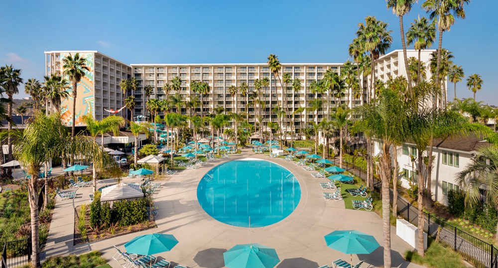 A large circular swimming pool surrounded by lounge chairs, umbrellas, palm trees, and a multi-story hotel in the background.