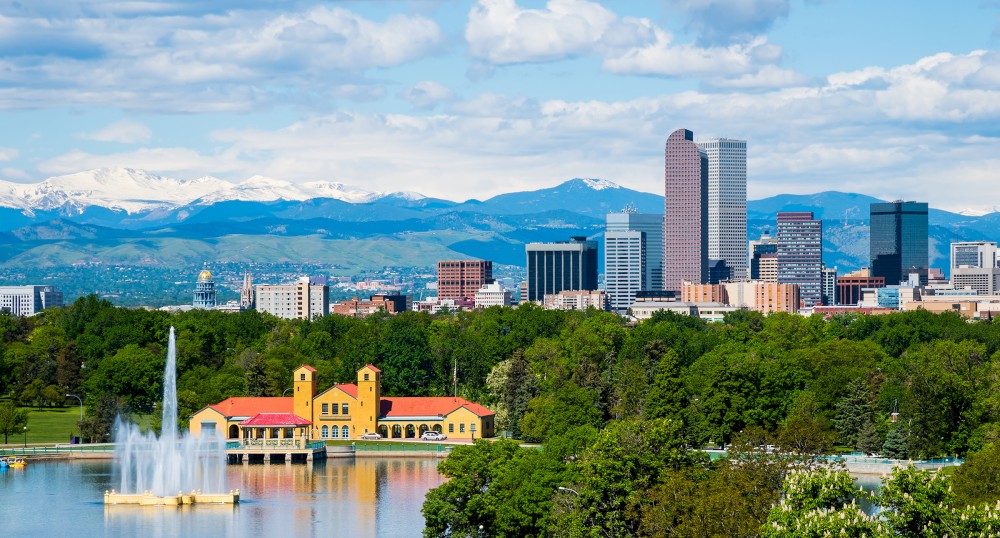 A scenic view of a city skyline with tall buildings, a yellow waterfront building, a pond with fountains, and mountains in the background ending the sentence.