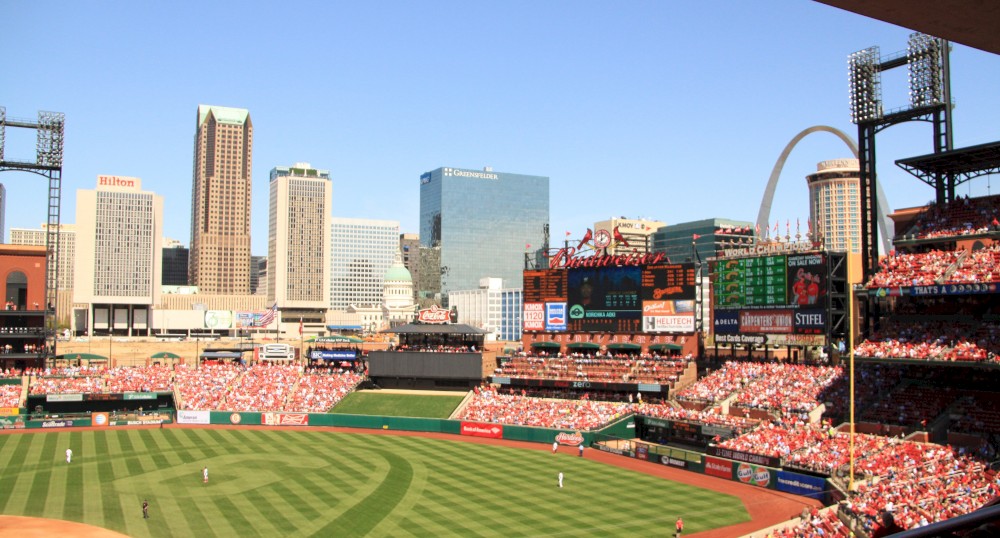 A bustling baseball stadium filled with fans, a city skyline, and an iconic arch are visible in the background, showcasing a lively game day.