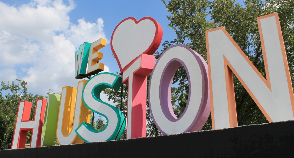 A colorful sign reads "WE ♥ HOUSTON" against a backdrop of trees and a partly cloudy sky.