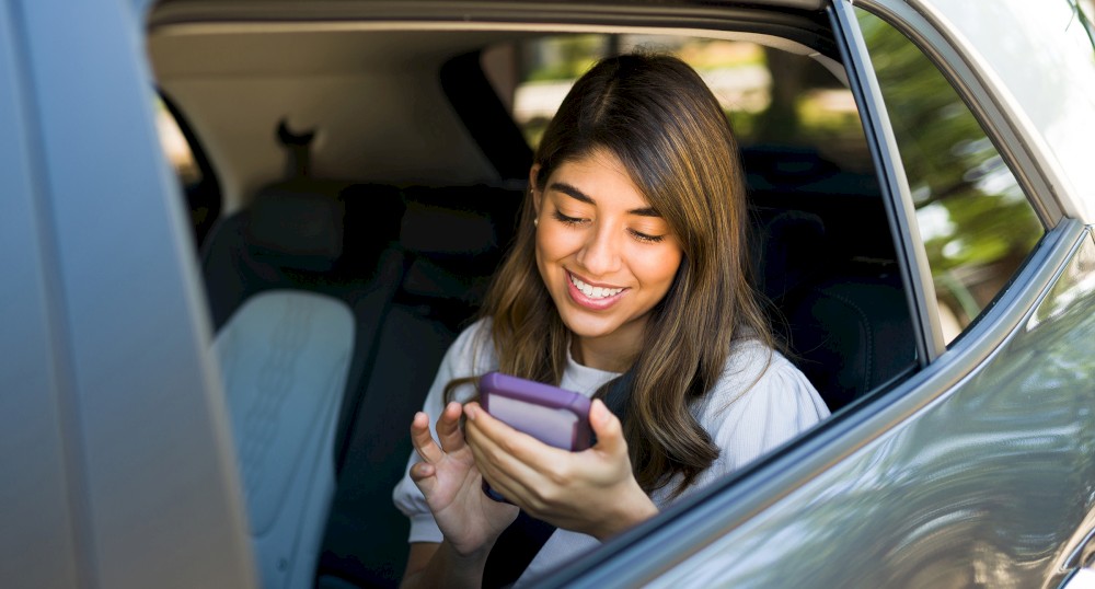 A person is sitting in a car, looking at a smartphone and smiling. The car window is open, and the person appears to be enjoying their device.