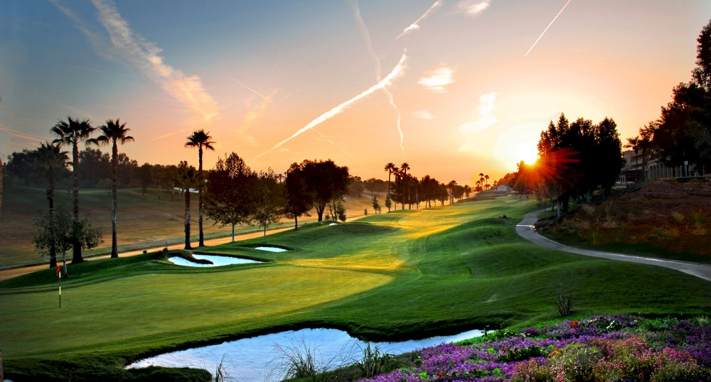 A scenic golf course at sunset, featuring palm trees, vibrant flowers, a water hazard, and sand bunkers, with the sun setting in the background.