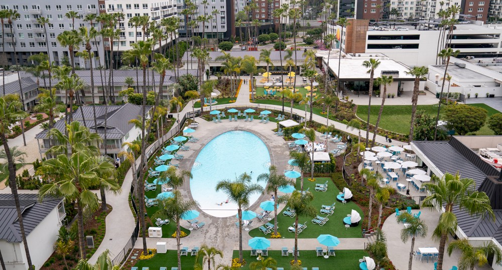 An aerial view of a residential complex with a large oval swimming pool, lounge chairs, tables with umbrellas, multi-story buildings, and greenery.