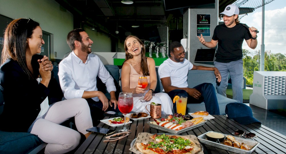 A group of five people enjoying drinks and food, seated and standing around a table with pizza and other dishes in an outdoor setting.