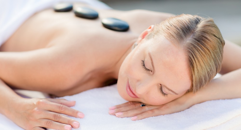 A person is lying face down, relaxing with hot stones placed on their back, covered partially with a towel.