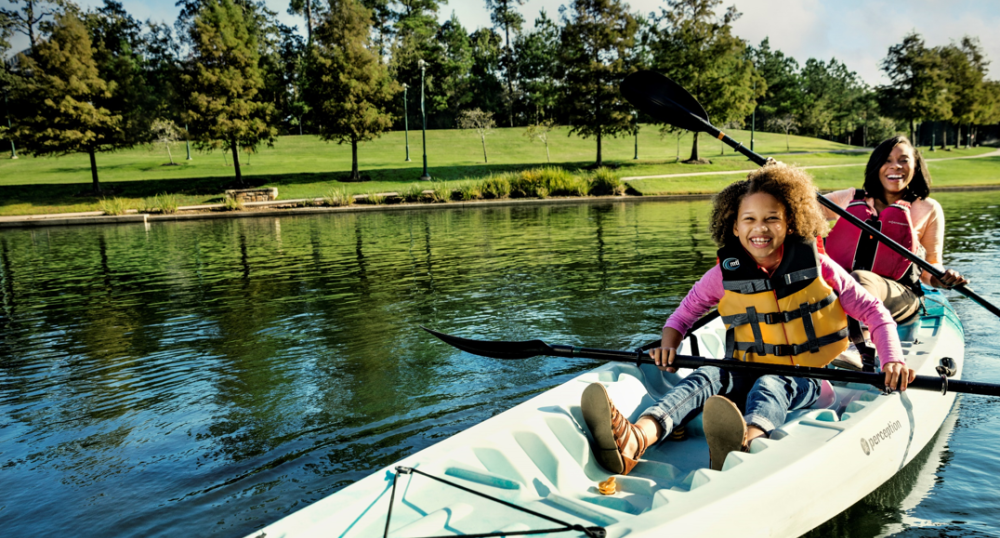 Two people are enjoying a sunny day in a kayak on a serene body of water, with trees and greenery in the background.