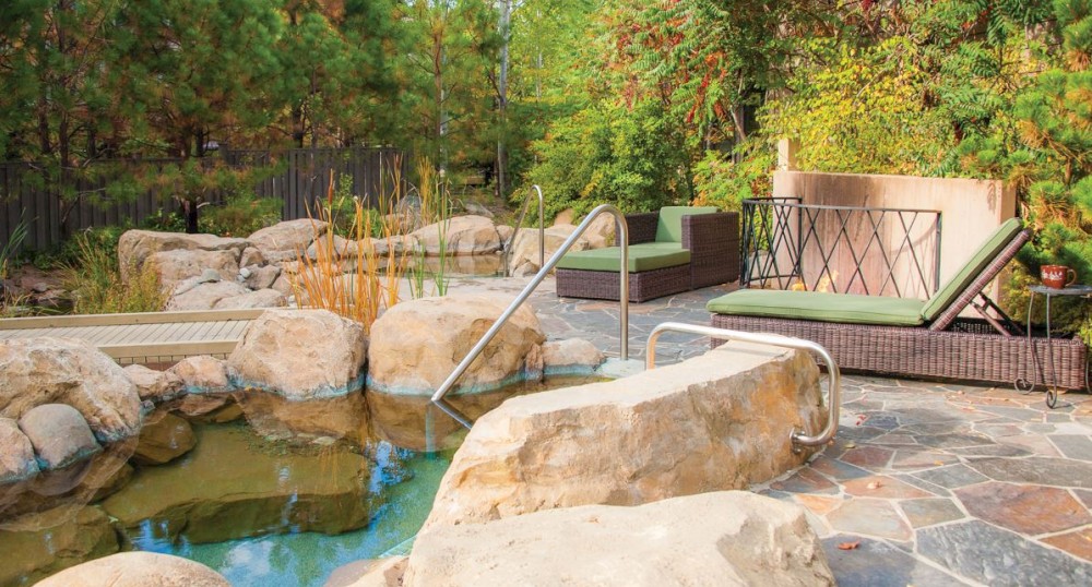 An outdoor area featuring a stone-lined hot tub, loungers with green cushions, and surrounded by lush greenery and rocks ending the sentence.