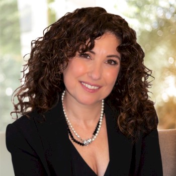 A woman with curly hair is smiling at the camera, wearing a black outfit and a pearl necklace, against a softly blurred background.
