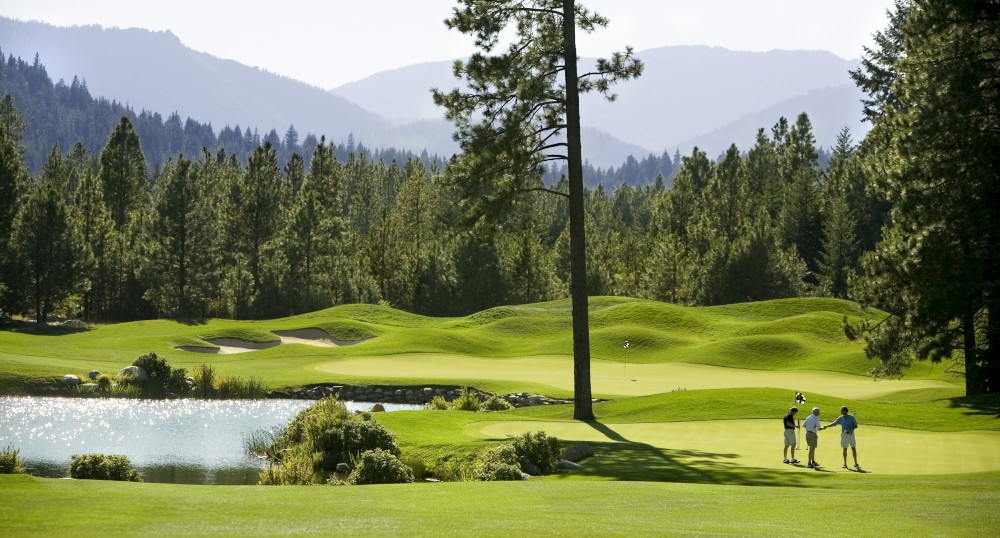 A picturesque golf course with lush green fairways, a pond, and surrounding mountains. Three individuals are seen enjoying a day of golf.