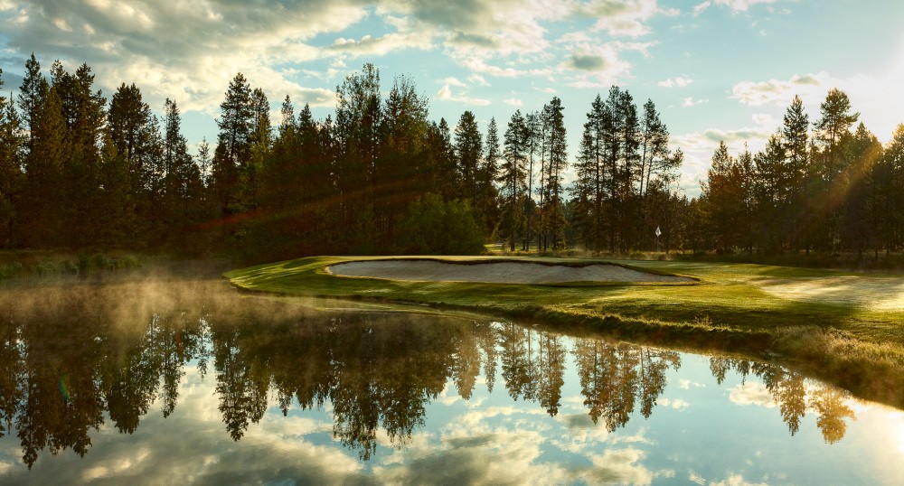 The image shows a serene golf course with a sand trap and lush greenery, complemented by a reflective water hazard under a partly cloudy sky at sunset.