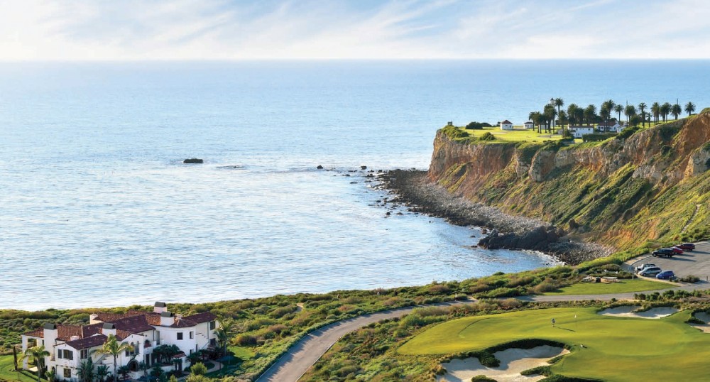 This image shows a coastal landscape with a building near a golf course, a cliff, and the ocean under a partly cloudy sky in the background.
