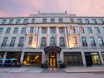 A multi-story building with tall columns, flags, multiple lit windows, and an arched entrance, photographed at dusk from street level.