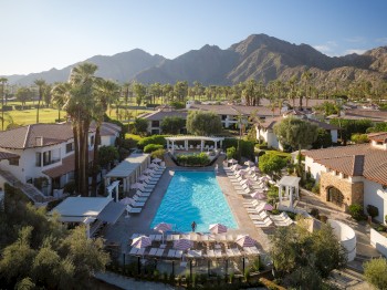 A luxurious resort with a central swimming pool, surrounded by cabanas and loungers, nestled against a backdrop of mountains and palm trees.