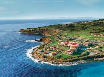An aerial view of a coastal resort with buildings, green landscape, and cliffs next to the ocean, with the horizon visible in the background.
