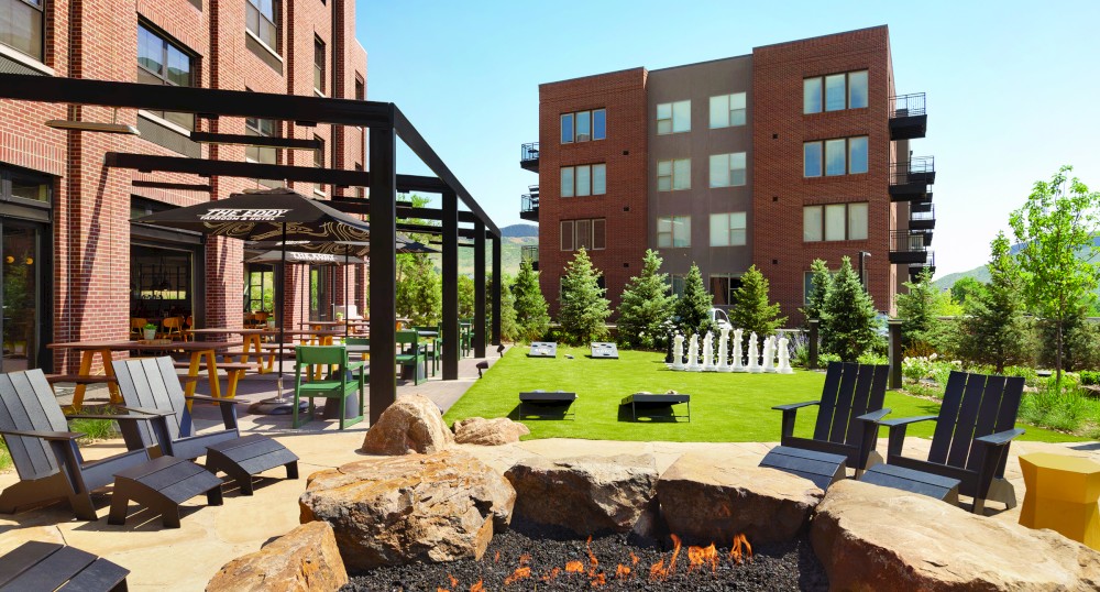 The image shows an outdoor communal area with a fire pit, seating, greenery, and oversized outdoor chess set, surrounded by apartment buildings.