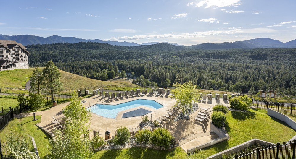 This image shows a hexagonal swimming pool surrounded by lounge chairs, set in a scenic mountain landscape under a sunny sky.