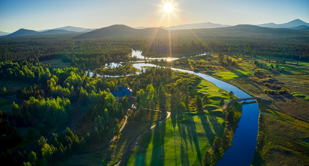 An aerial view of a river meandering through a lush green landscape with mountains in the background and the sun setting in the sky.