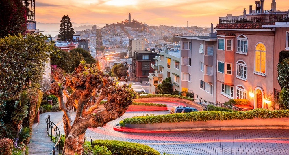 A winding, steep street lined with houses, lush greenery, and a view of a hilly cityscape at sunset, featuring a large landmark in the distance.