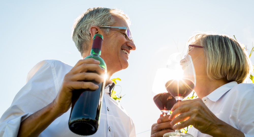 Two people are smiling, holding glasses of red wine and a bottle of wine, with clear skies in the background.