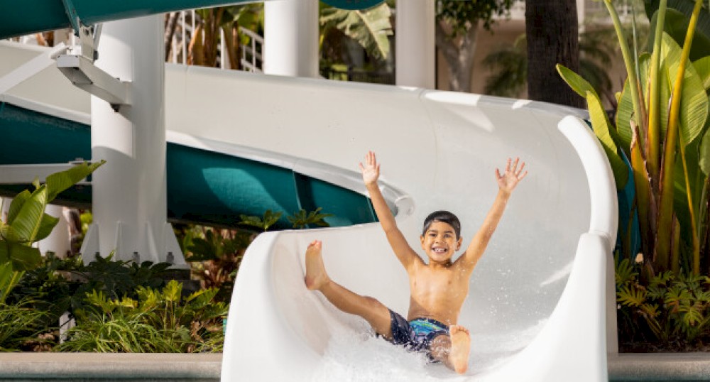 A child is joyfully sliding down a white water slide into a pool, with the large green "Twister" slide visible in the background.