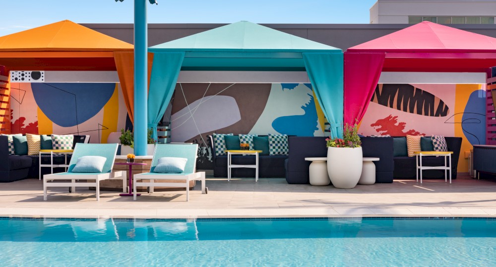 Poolside cabanas with colorful awnings and lounge chairs by a blue pool, featuring vibrant wall art and white planters.