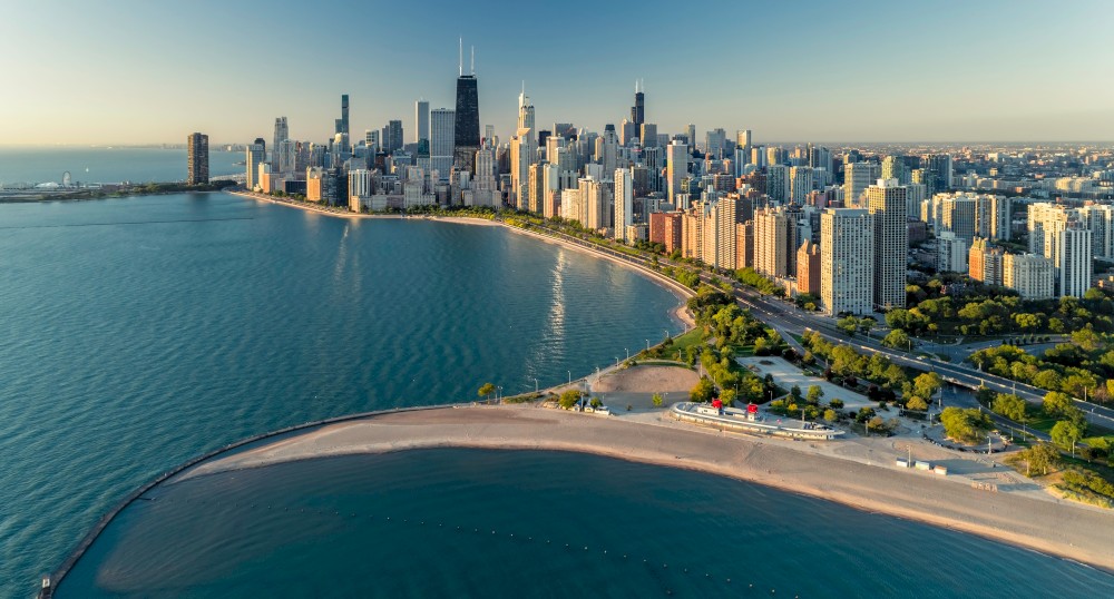 Aerial view of a city skyline beside a large body of water, with a curved beach and greenery in the foreground.