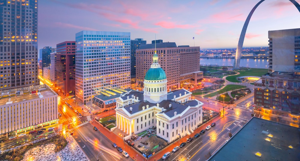 A cityscape featuring a historic courthouse, modern buildings, and a large arch structure in the background during twilight, ending the sentence.