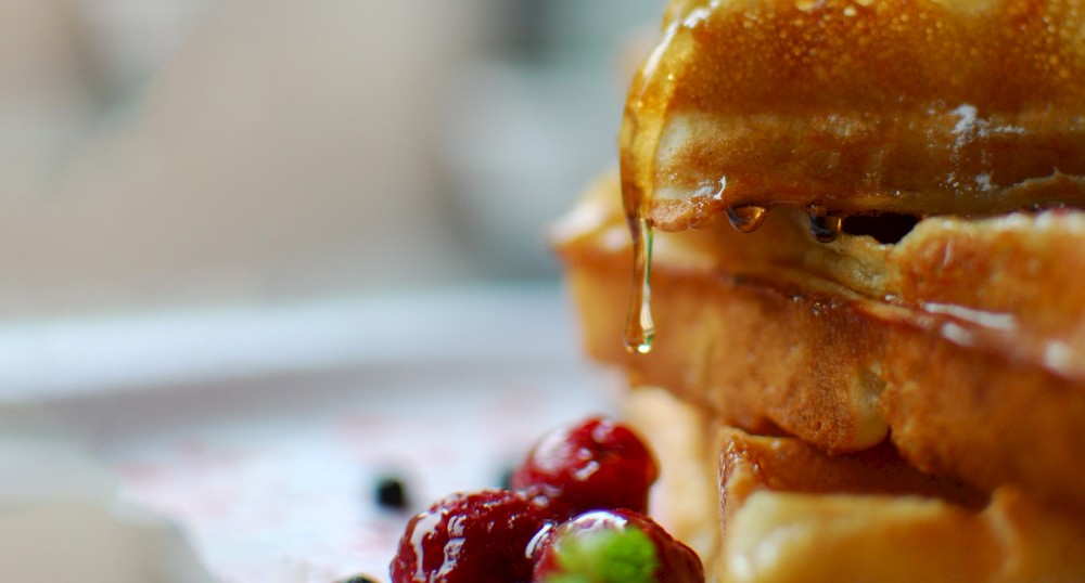 The image shows waffles being drizzled with honey or syrup, accompanied by fresh berries and greenery.