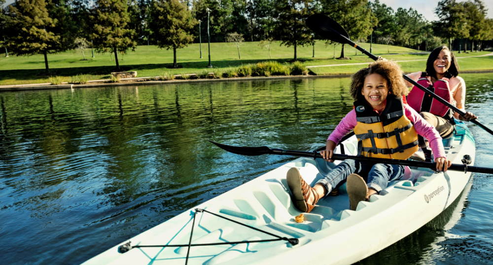 Two people are kayaking on a calm lake with life jackets on, enjoying the sunny day. Trees and grassy banks are in the background.