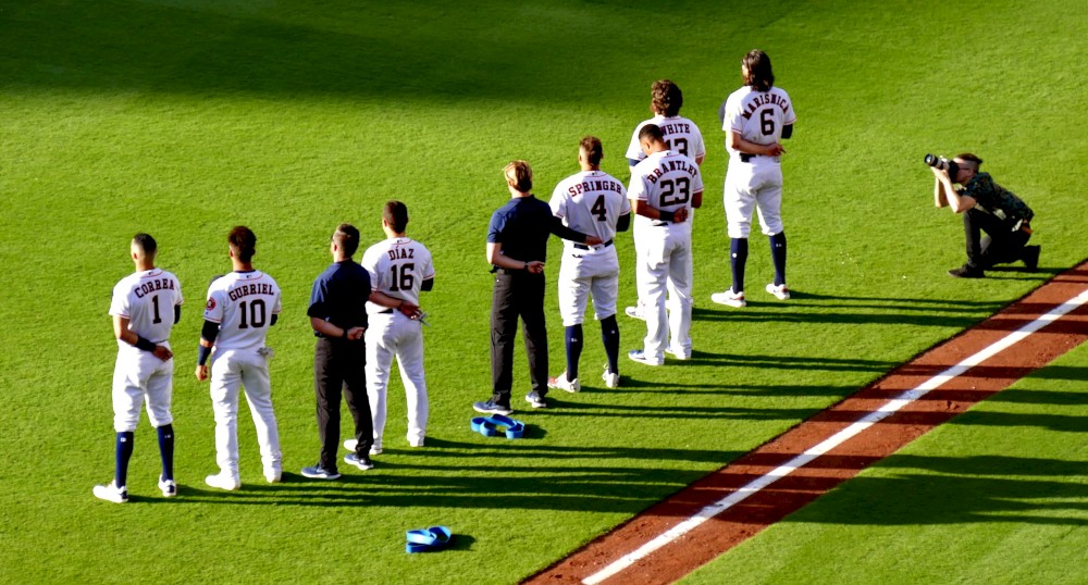 A group of baseball players are standing on the field in a line, possibly during the national anthem, while a photographer captures the moment.