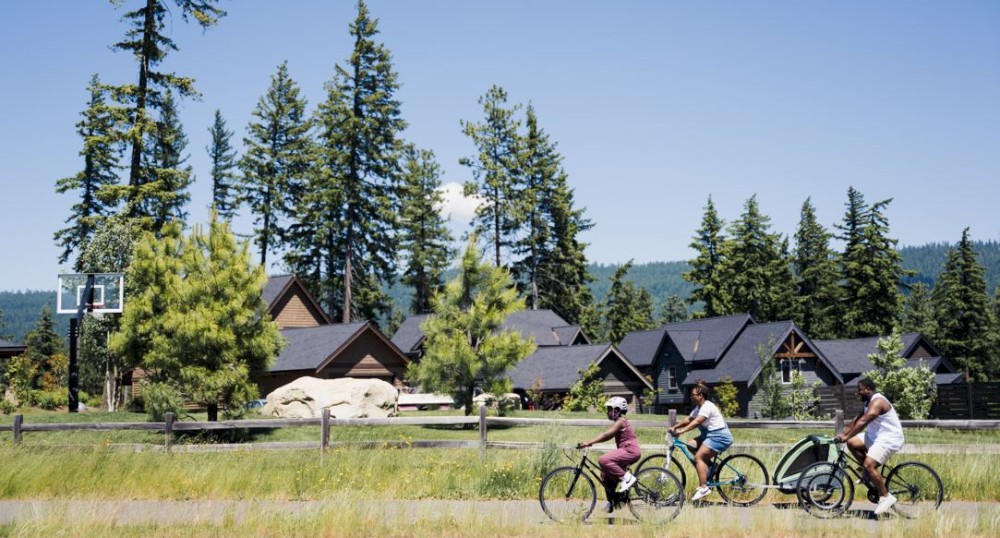 Three people are riding bicycles on a paved path with houses and tall trees in the background under a clear blue sky.