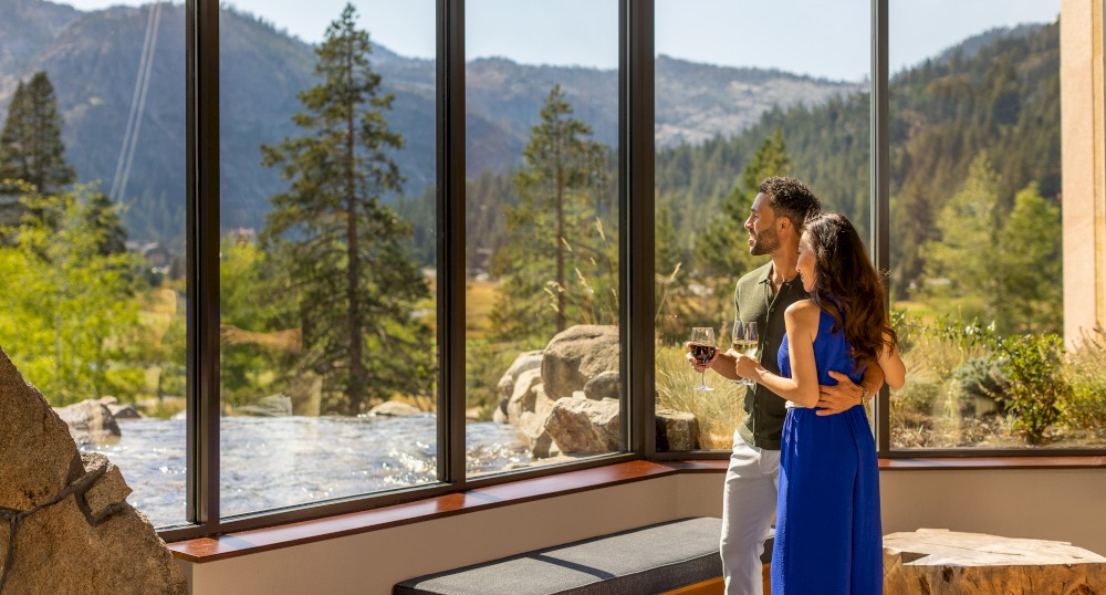 A couple stands by large windows, overlooking a scenic mountain and forest view. They are dressed casually, with the woman in a blue dress.