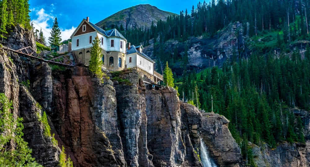 A picturesque house atop a cliff with a mountain and lush forest in the background and a waterfall flowing down the cliff face.