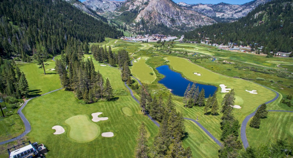 This image features a scenic golf course surrounded by trees and mountains, with a body of water in the middle. The sky is clear and blue.