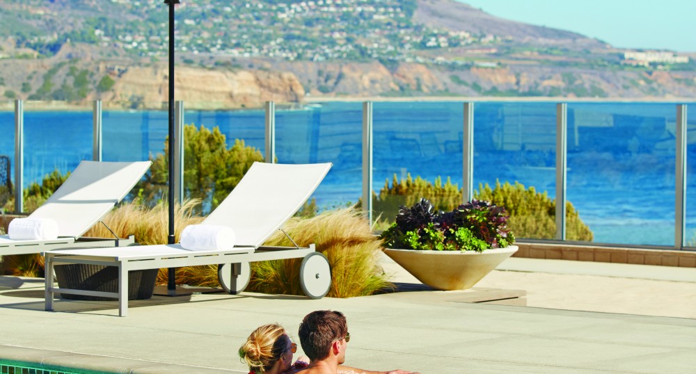 A couple embraces in a pool with a scenic ocean view, lounge chairs, and an umbrella on the deck.