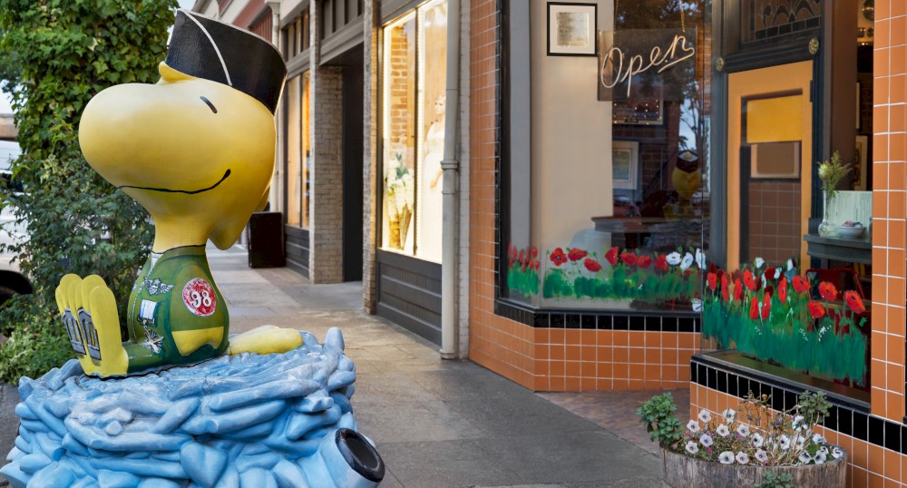 A statue of Snoopy dressed in a military uniform is in front of a burger restaurant with a neon "Open" sign and flower decorations in the window.