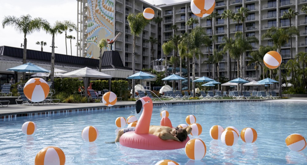 A man relaxes on a pink flamingo float in a hotel pool surrounded by numerous orange and white beach balls, with palm trees and a building nearby.