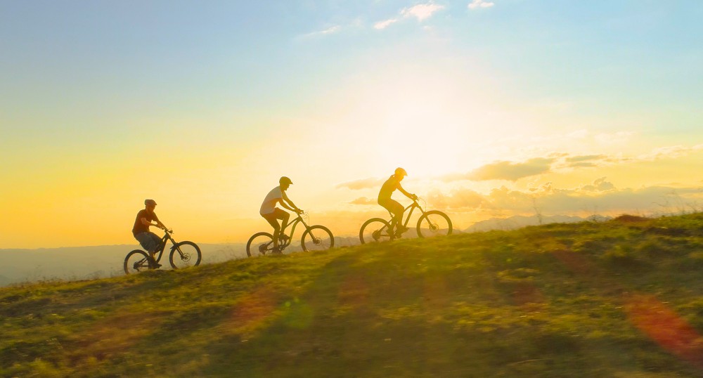 Three people are riding mountain bikes on a grassy hill during sunset, with the sky glowing in the background.