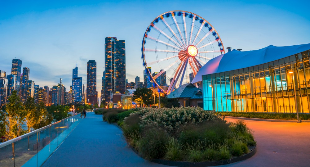 The image features a cityscape during twilight with a Ferris wheel, modern buildings, and a lit-up pavilion along a pedestrian pathway.