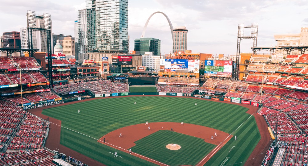 A baseball stadium with a game in progress, surrounded by city buildings and an iconic arch in the background. Fans are seated in the stands.