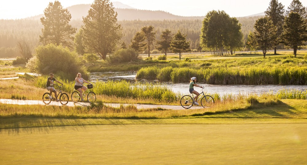 Three cyclists ride along a pathway beside a river surrounded by lush greenery and trees, with mountains in the background and sunlight filtering through them.