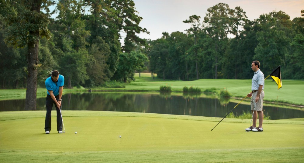 Two people are on a golf course. One is putting on the green, while the other stands nearby holding the flag. Trees and a pond are in the background.