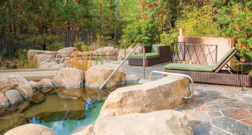 A serene outdoor area featuring a pond with rocks and a handrail, surrounded by trees and seating with green cushions.